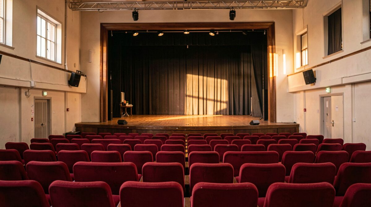 An empty small town theater auditorium with red seats and a lit stage, photographed from the back row in warm late afternoon light, wide framing.